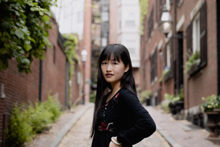 Rebecca F Kuang standing outside in front of an apartment block, wearing black, looking at the camera
