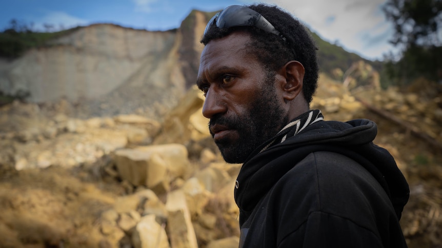 A young Papua New Guinea man wearing a black hoodie and sunglasses looks sombrely out over a large pile of rocks.