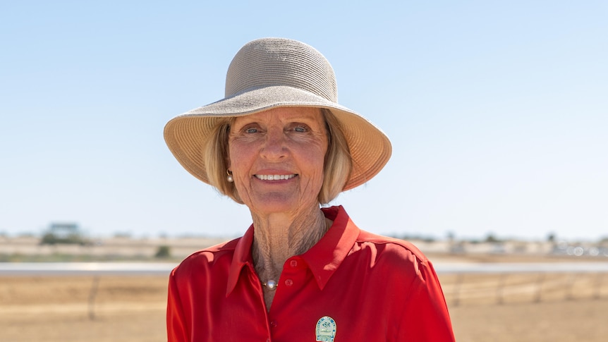 A lady in a red shirt and green pants with a hat on and smiling on the Birdsville race track
