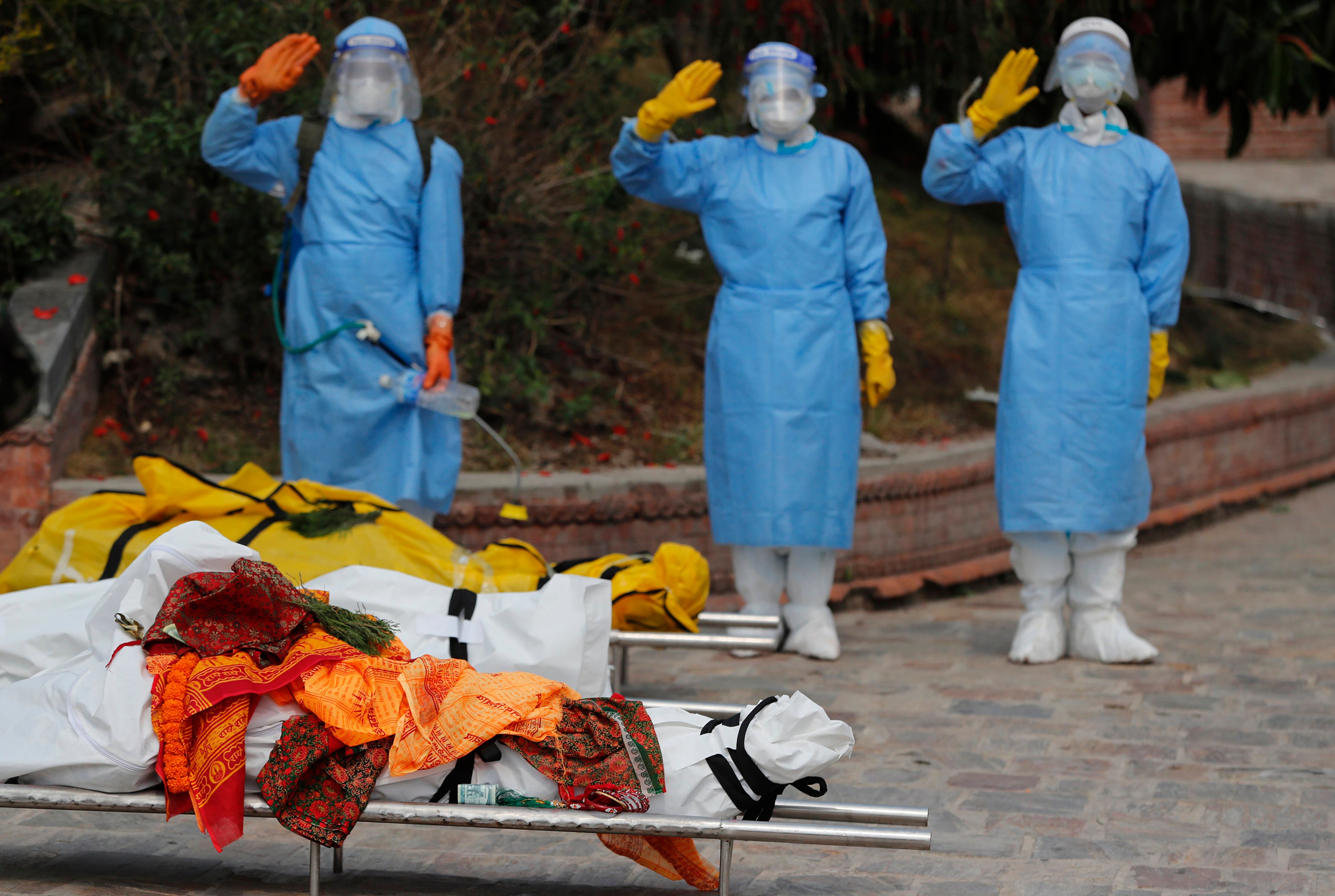 Three people in full PPE salute behind two stretchers carrying bodies wrapped in fabric.