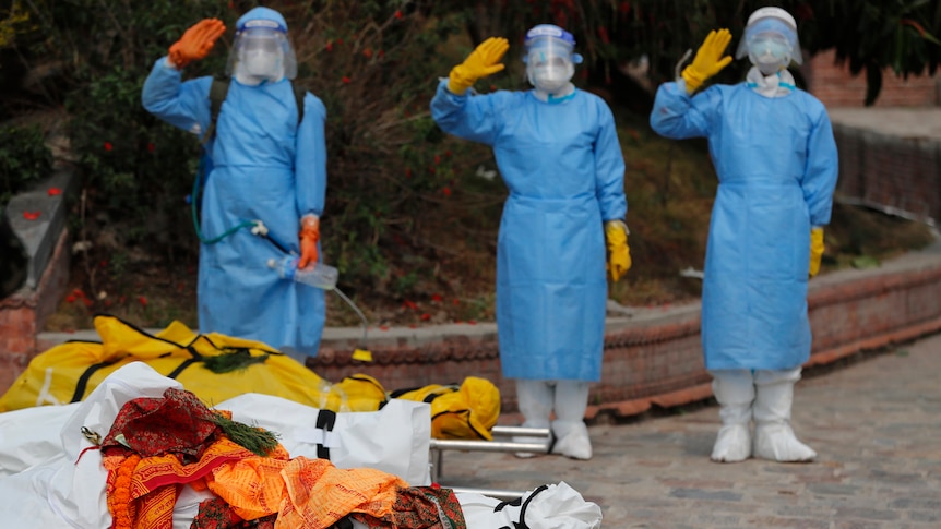 Three people in full PPE salute behind two stretchers carrying bodies wrapped in fabric.