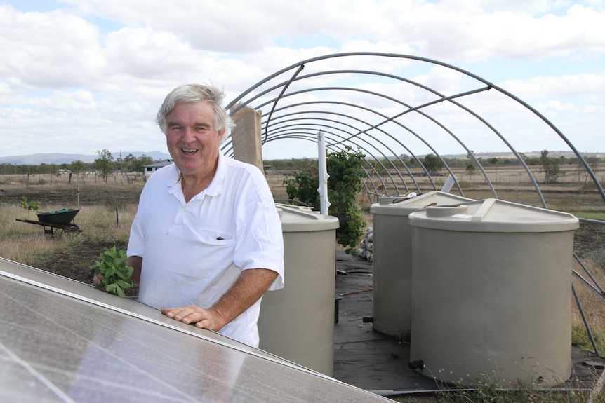 An older man with his hands on a solar panel, standing in front of an aquaponics set-up.