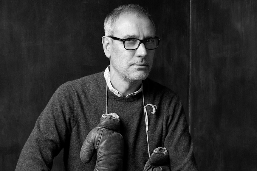 A black and white photograph of a man with glasses with boxing gloves over his shoulders.