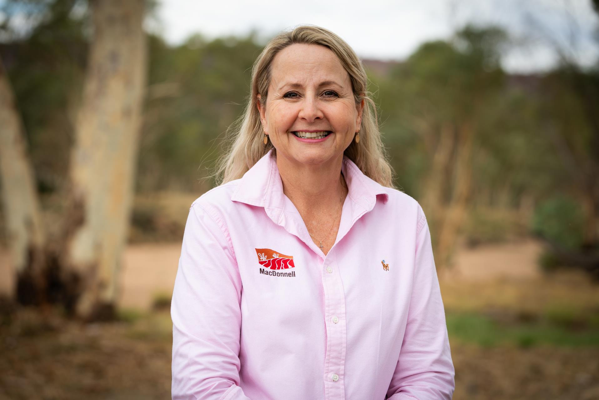 Belinda Urquhart, with blonde hair and wearing a pink shirt, smiling. The photo is taken outside.