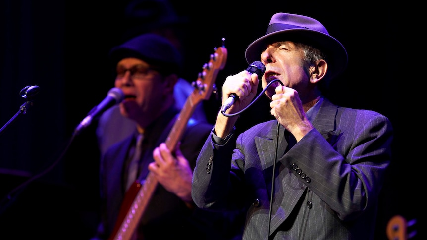 An older man in a suit and hat sings into a microphone on stage, standing on front of an out-of-focus bass player