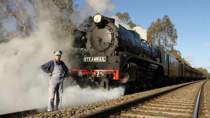 A man waring overalls and a cap standing on a trainline in front of a steam engine blowing smoke.