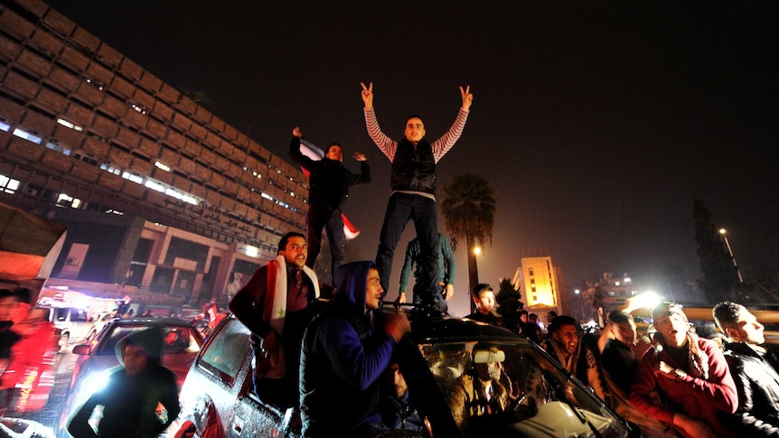 Men stand on top of a car and some sit on the windowsills celebrating in the rain