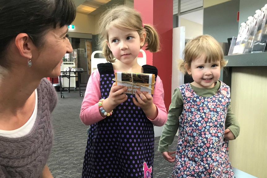 Two young girls hold a packet of seeds in a library as their mother looks on.