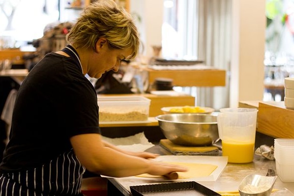 A chef leans over a commercial kitchen table, using a rolling pin to flatten out a pastry to turn into a lemon tart.