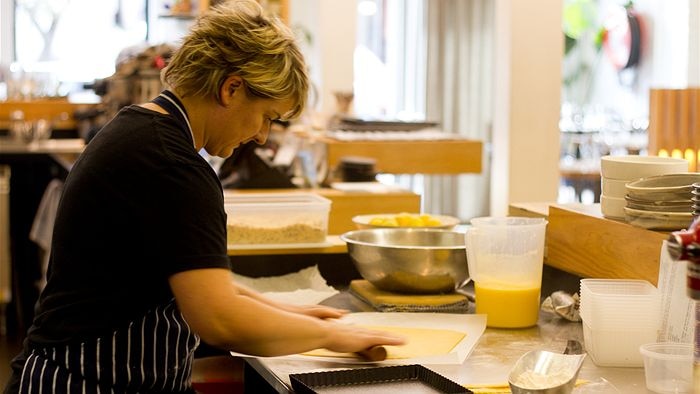 A chef leans over a commercial kitchen table, using a rolling pin to flatten out a pastry to turn into a lemon tart.