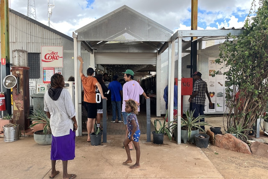 A group of people are gathered at the entrance of a tin building. The sky is cloudy, with pockets of blue.