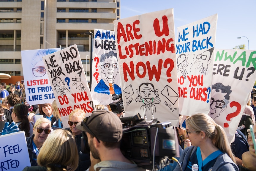 Nurses and doctors rallying outside a hospital, holding a number of signs 