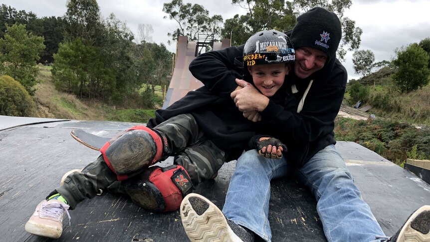 A father and son sitting at the top of a giant skate ramp, hugging.