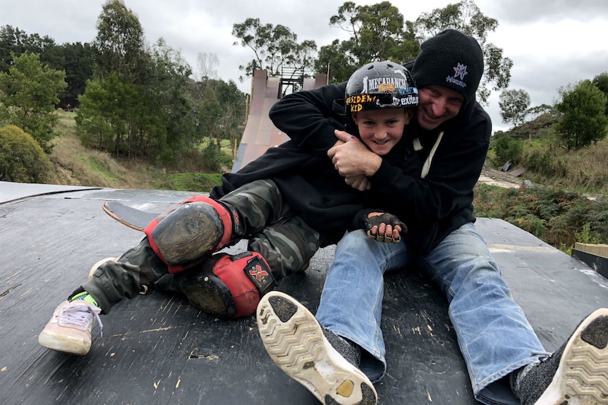 A father and son sitting at the top of a giant skate ramp, hugging.