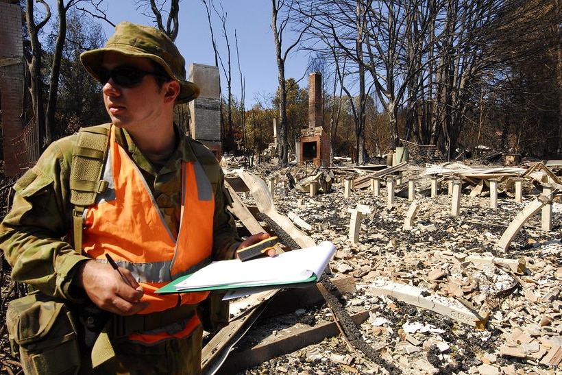 Search Task Group's Private Adam Honeybun writing in clipboard with destroyed building behind him.