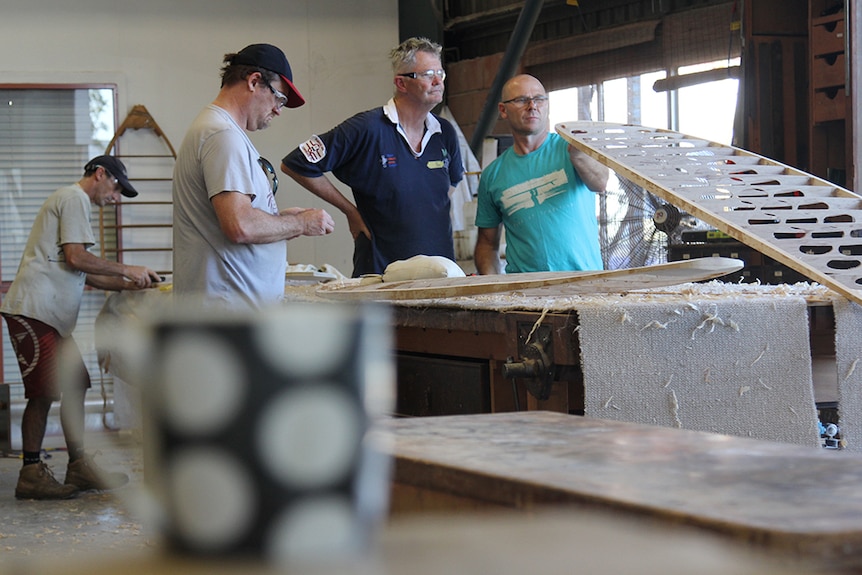 Two men inspect the curve of a surfboard frame, a black and white mug in the foreground.