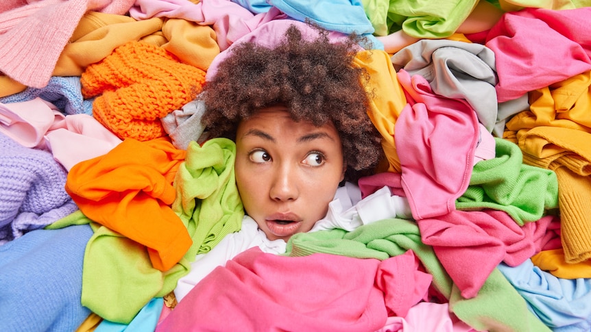 A woman with a surprised expression pokes her head out of a pile of brightly coloured clothes