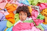 A woman with a surprised expression pokes her head out of a pile of brightly coloured clothes