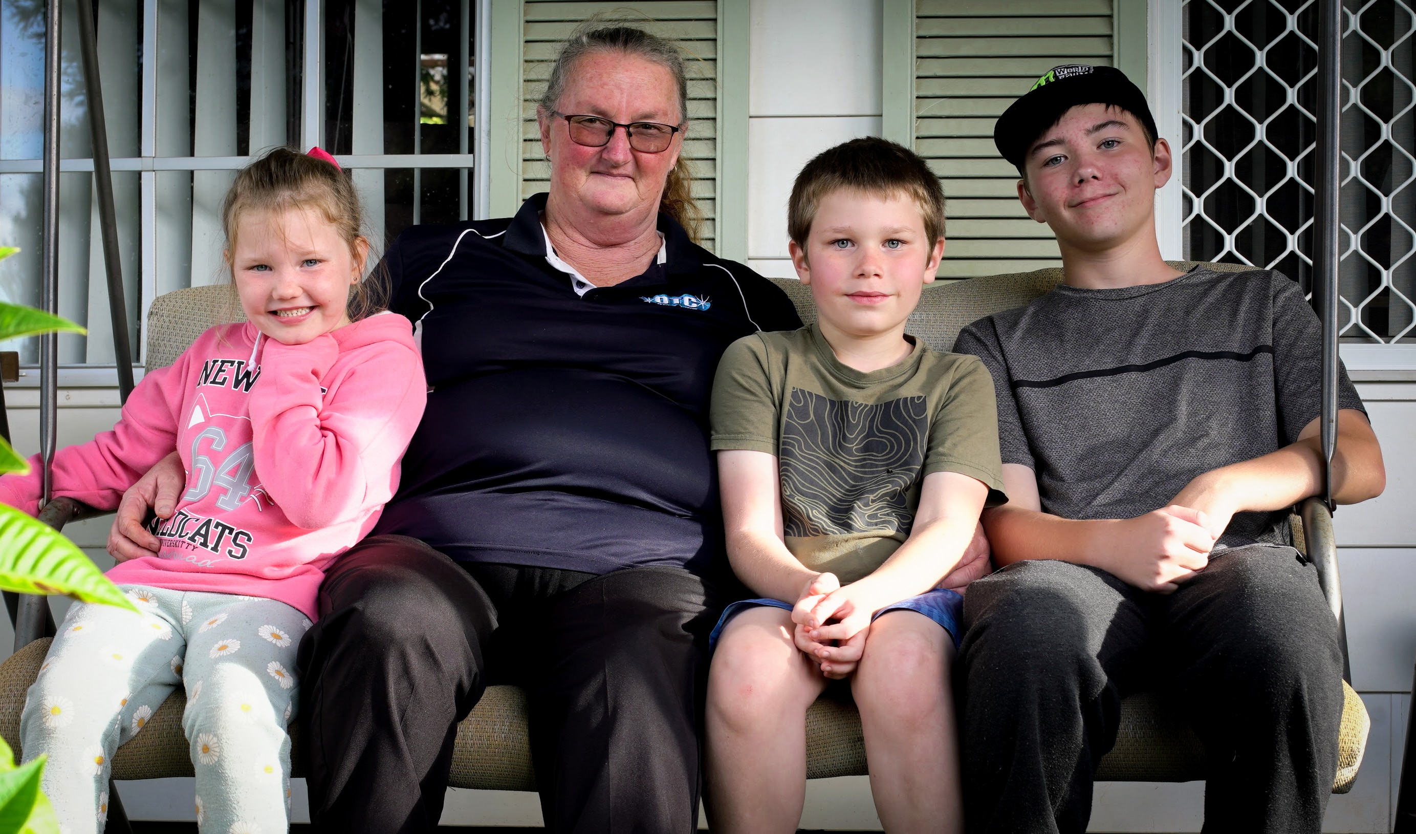 Raeleen sits with her three grandkids - a little girl wearing pink and two boys in T-shirts - out the front of their house.