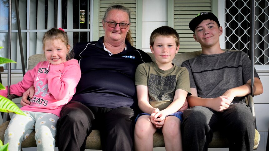 Raeleen sits with her three grandkids - a little girl wearing pink and two boys in T-shirts - out the front of their house.