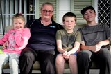Raeleen sits with her three grandkids - a little girl wearing pink and two boys in T-shirts - out the front of their house.