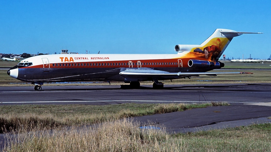 Colour image of ‘TAA Central Australian’ plane on apron. Deep ochre stripe with Uluru painted on the tail.