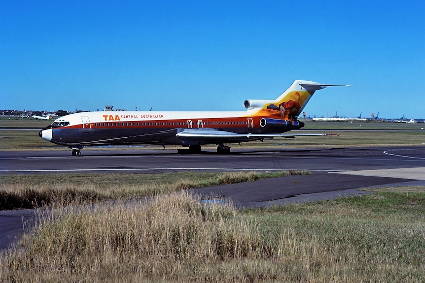 Colour image of ‘TAA Central Australian’ plane on apron. Deep ochre stripe with Uluru painted on the tail.