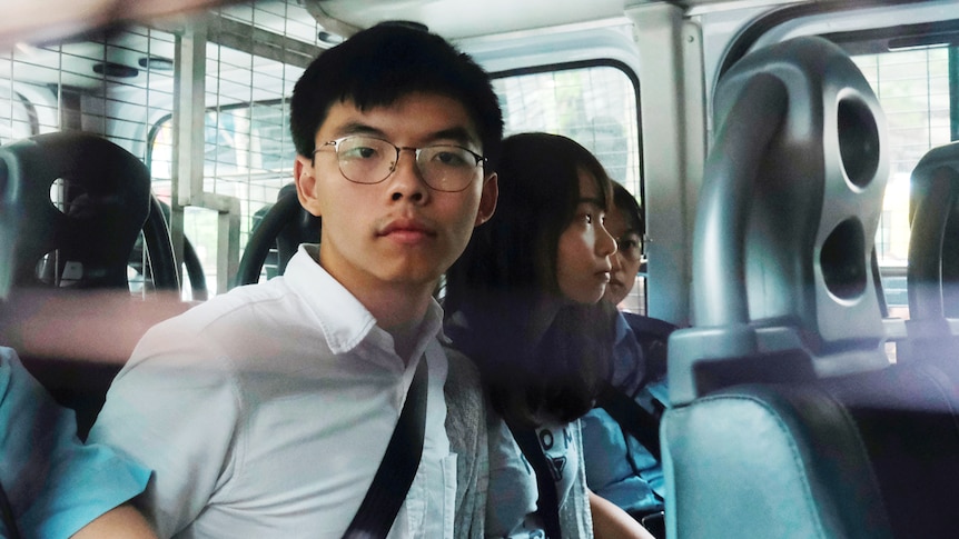 You see the back seat of a white police van carrying three young Hongkongers handcuffed with Joshua Wong at their centre.