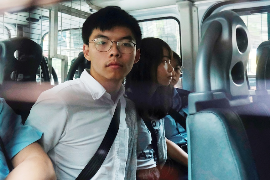 You see the back seat of a white police van carrying three young Hongkongers handcuffed with Joshua Wong at their centre.