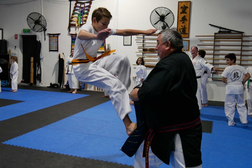 A male martial arts students leaping into the air and kicking a foam pad being held by an instructor.