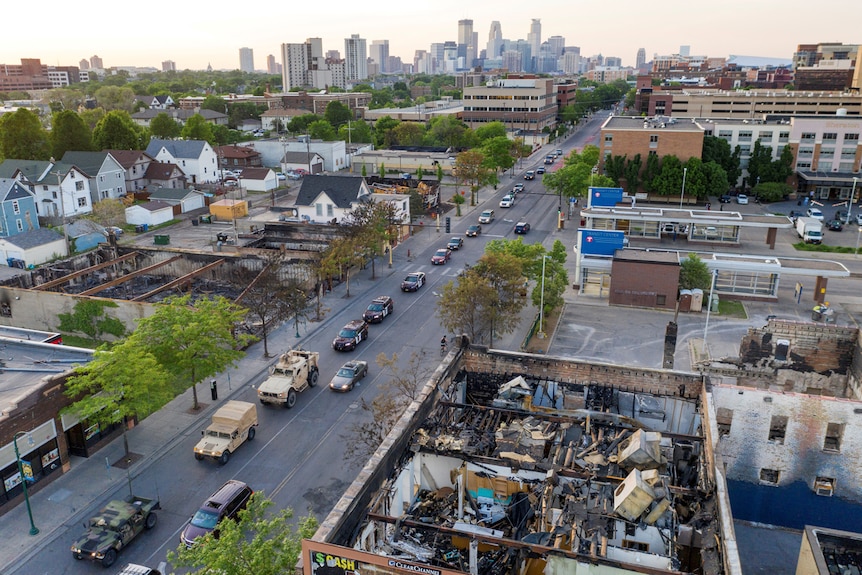 An aerial photo shows a long convoy of US military vehicles driving down a street past charred buildings in daylight.