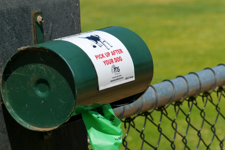 A poo bag dispenser bolted to a pole supporting a fence.