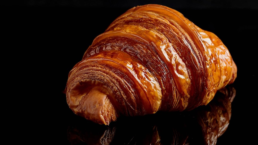 You view a crispy, golden croissant on top of a reflective, rich black surface. 