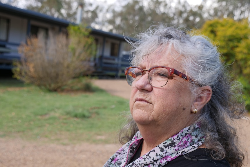 A grey-haired, bespectacled woman sits in a yard.