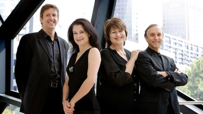 Four musicians in black standing in front of a window. City skyline in the background.