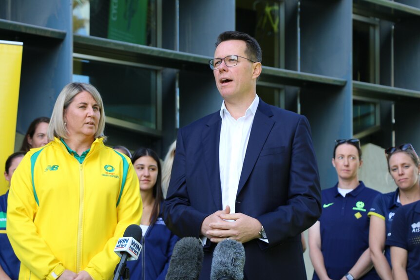 A man in a suit and a woman in a yellow jacket stand in front of microphones.