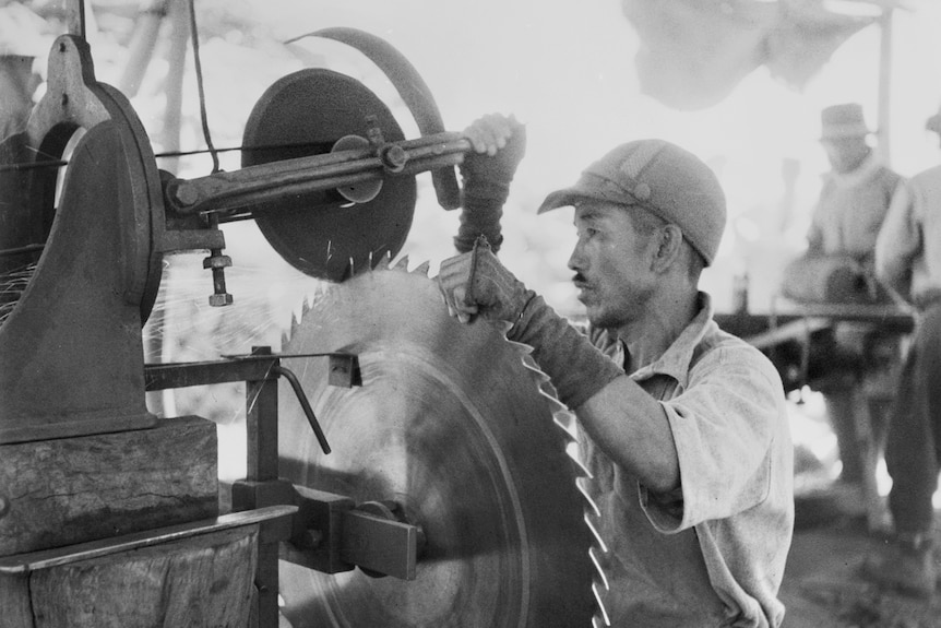 A man sits in front of a large disc saw, sharpening it with implements.