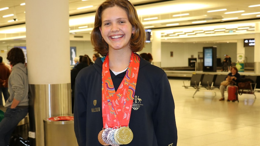 A young woman stands in an airport with seven medals on red ribbons around her neck.