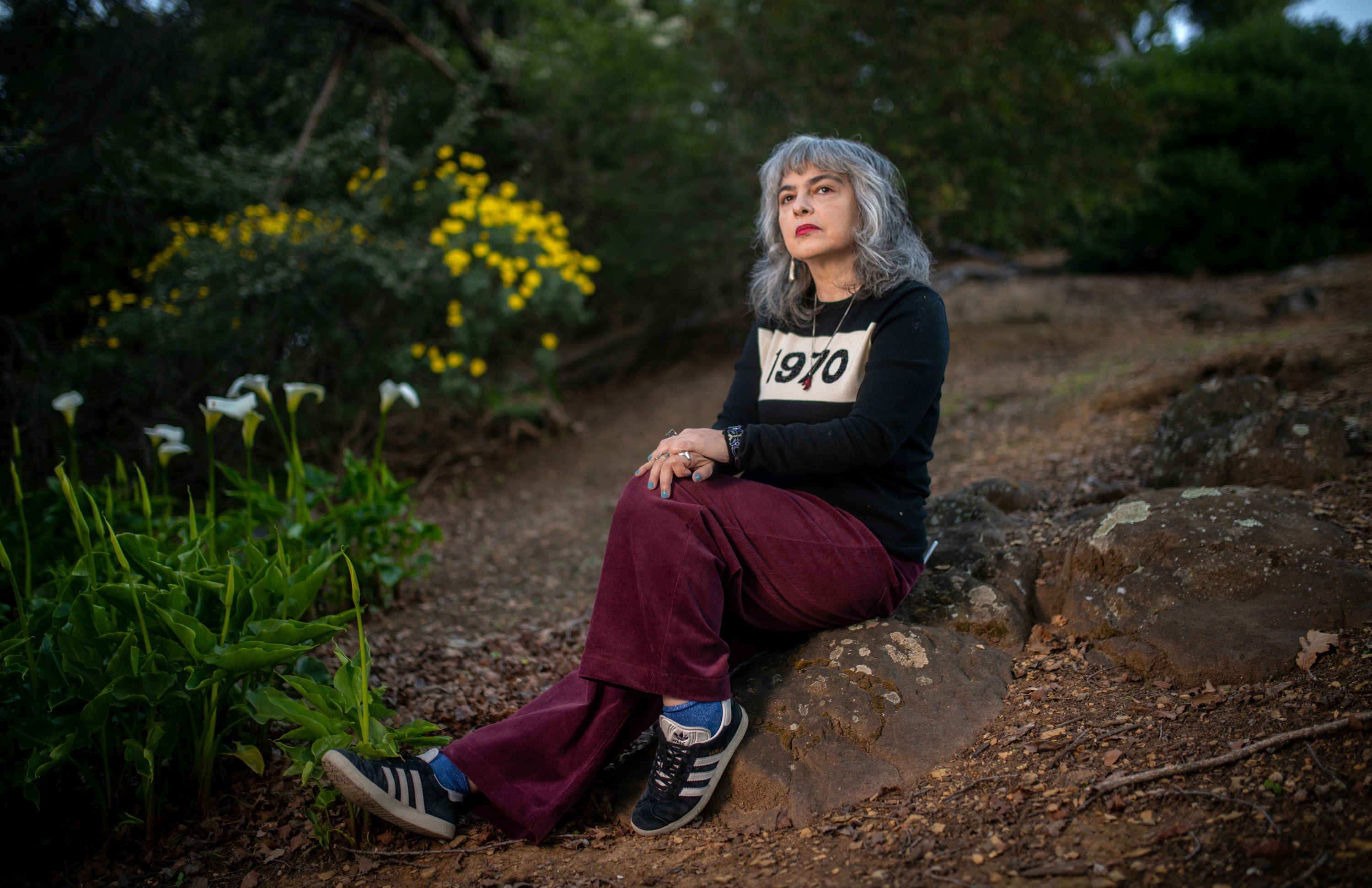 A woman sits on some rocks in a black jumper with "1970" printed and yellow and white flowers in the background.