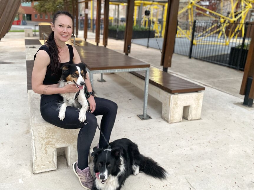 A woman wearing activewear sits at a bench with a dog on her lap and another at her feet