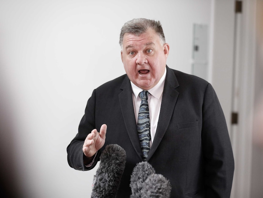 A white man with grey hair wearing a black suit and white shirt mid-speech with his mouth open and his hand out