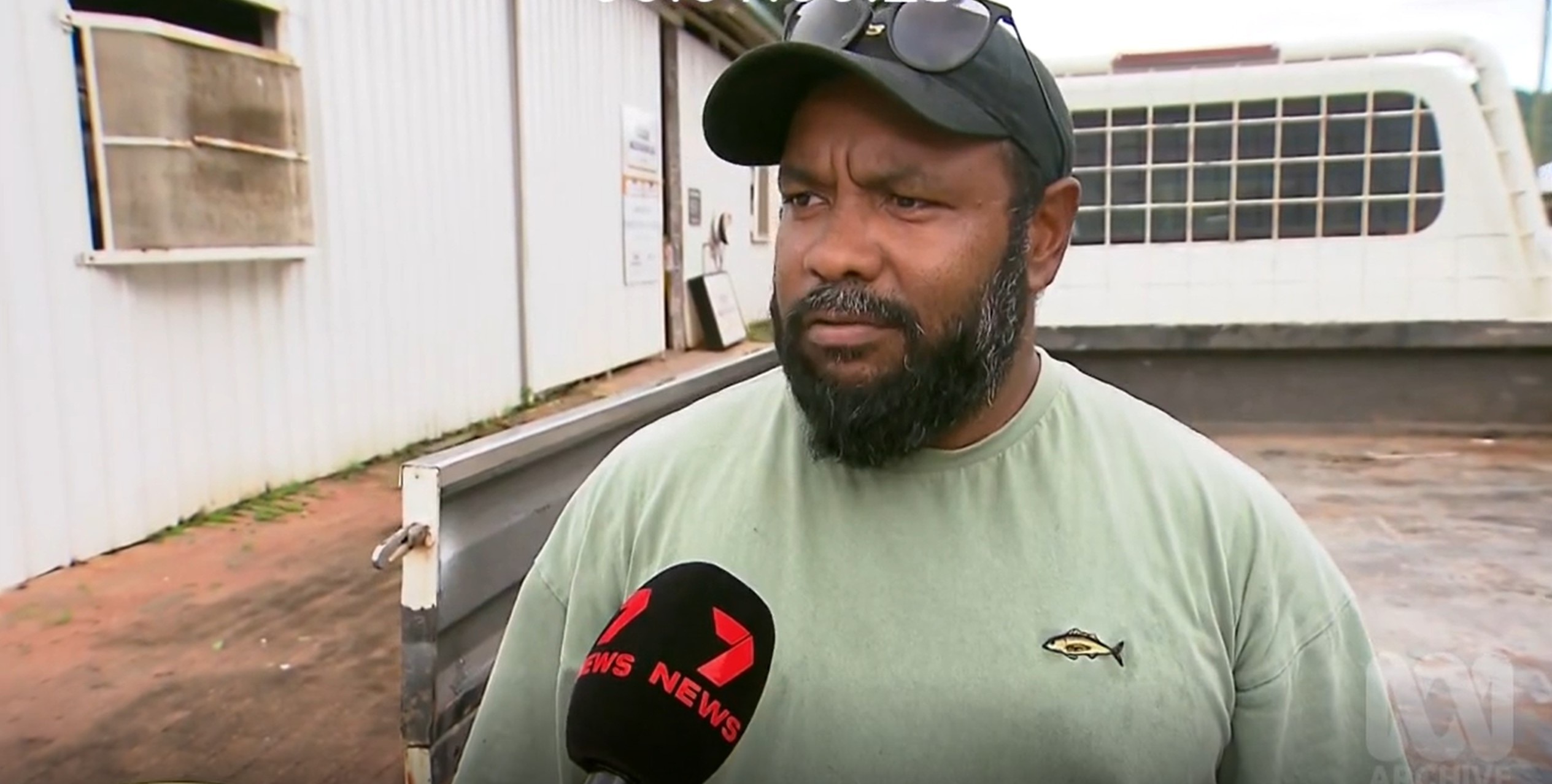 A man with a dark beard wears a cap while he stands in front of a building and speaks to a reporter.