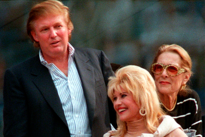 A man in a dark suit jacket and blue striped shirt stands behind a smiling blonde woman and an older woman in sunglasses