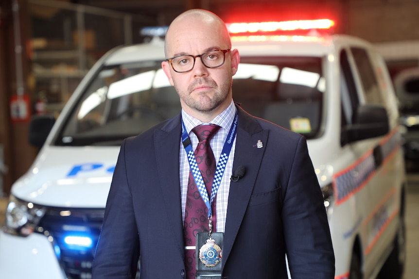 Bald man with glasses in suit and tie, with police badge on lanyard, in front of police van