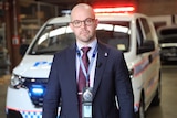 Bald man with glasses in suit and tie, with police badge on lanyard, in front of police van