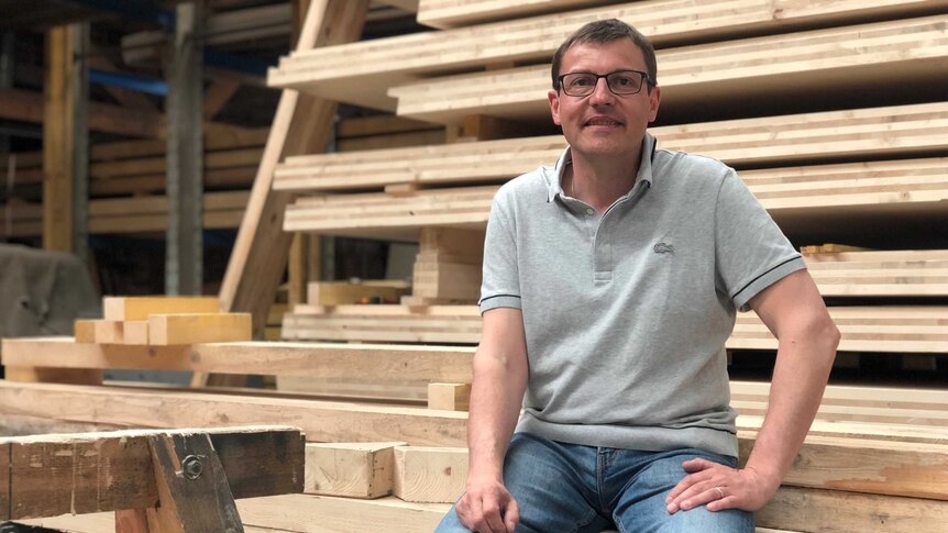 Daniel Flachat wears a grey Lacoste polo and blue washed jeans. He smiles as he sits among planks of wood.