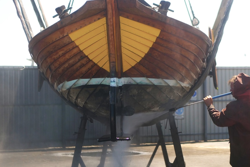 A man in red wet weather gear hoses down the hull of a large wooden boat with yellow bow on stand.
