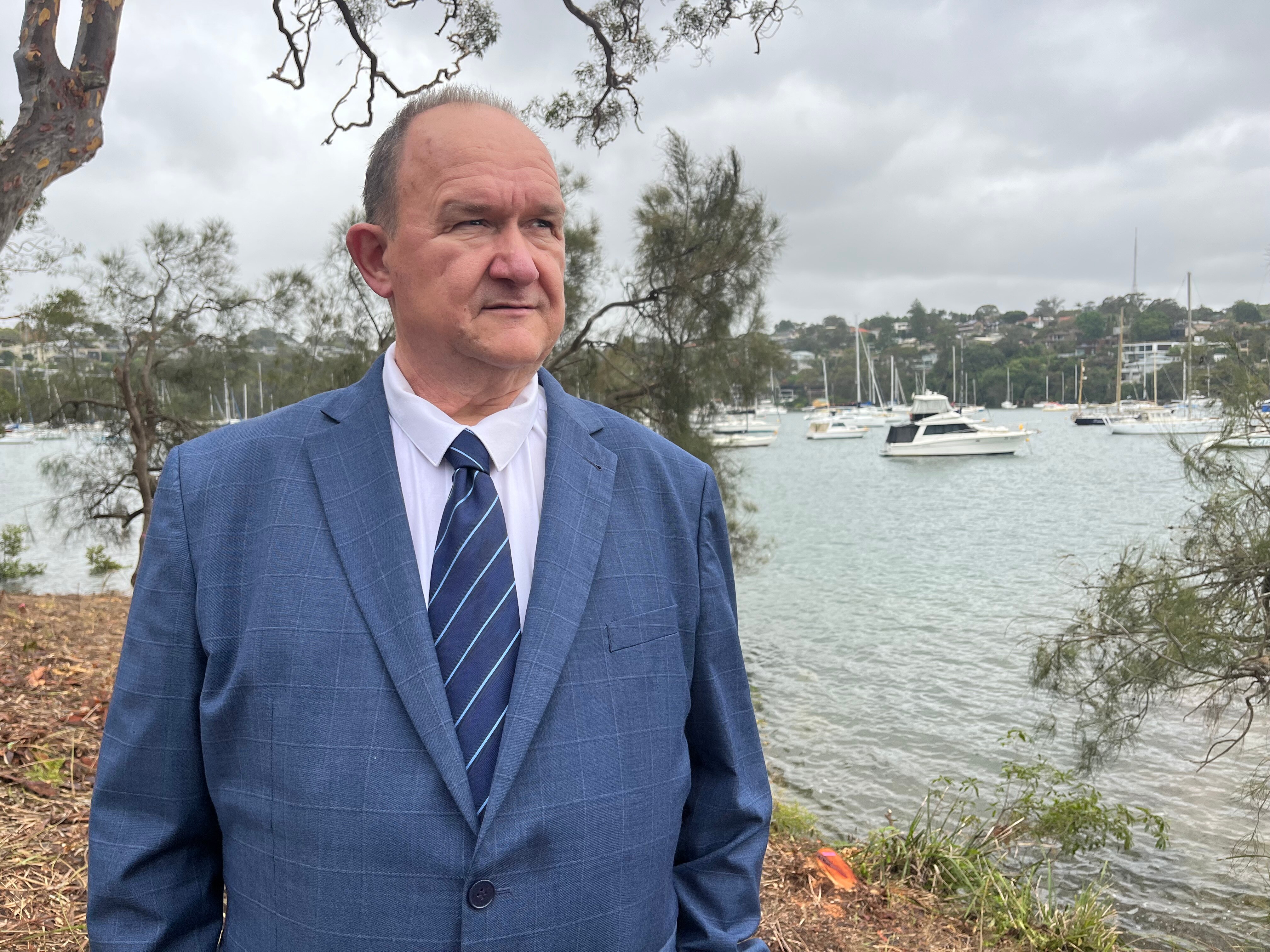 An elderly man in a suit stands by the water with boats in the background.