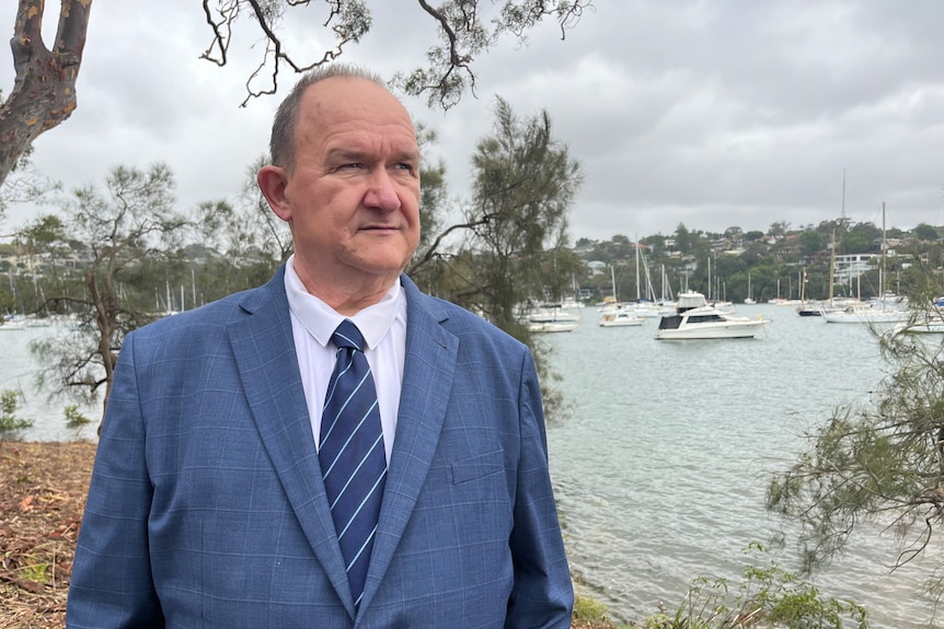 An elderly man in a suit stands by the water with boats in the background.
