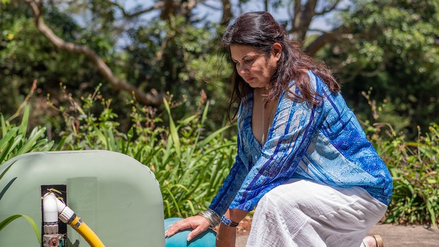A woman in a blue shirt and long white dress kneels before a water pump, with a concerned look on her face.